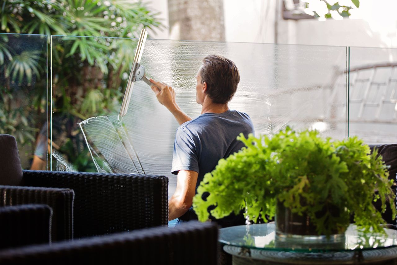 services-04 A man cleaning a glass barrier outdoors with a squeegee, surrounded by greenery.